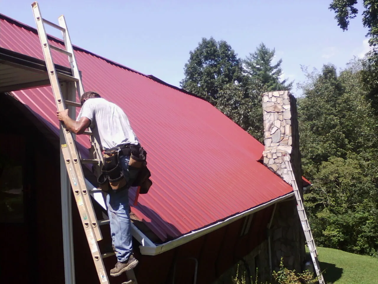 Roofer on a ladder beside a red metal roof and stone chimney — Soffit & Fascia Repair by Roofing Brothers in Aspinwall